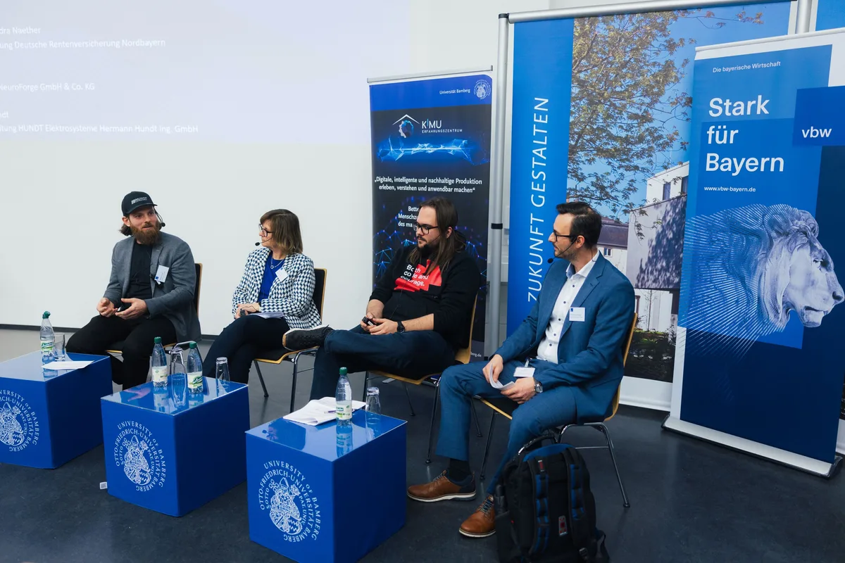 Podiumsrunde von schräg vorn, vier Personen vor Roll-ups, davor blaue Würfel mit Uni-Siegel und Wasserflaschen