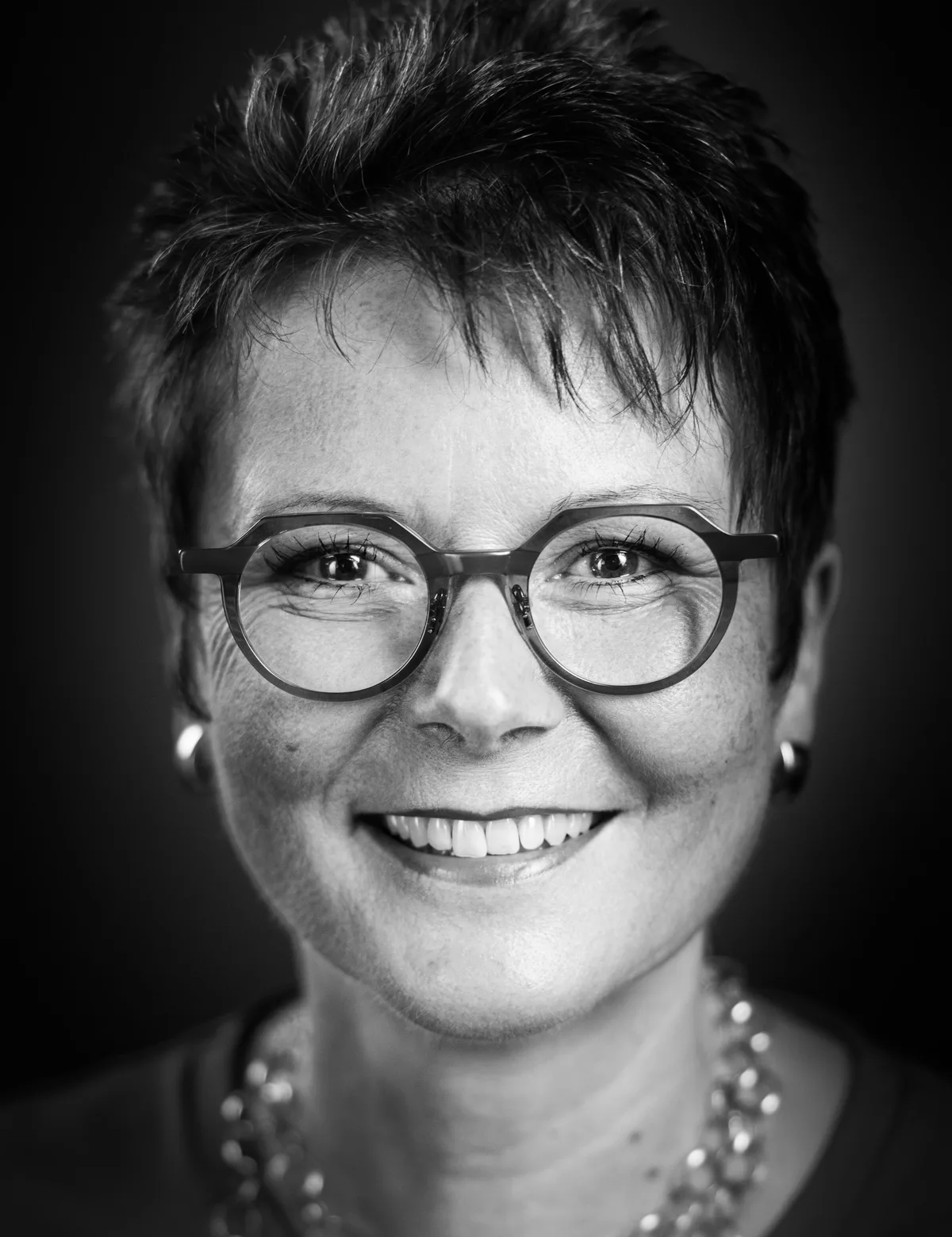 Lächelnde Frau mit kurzem dunklem Stufenhaar, runder Metallbrille und Perlenkette ganz nah am Gesicht fotografiert