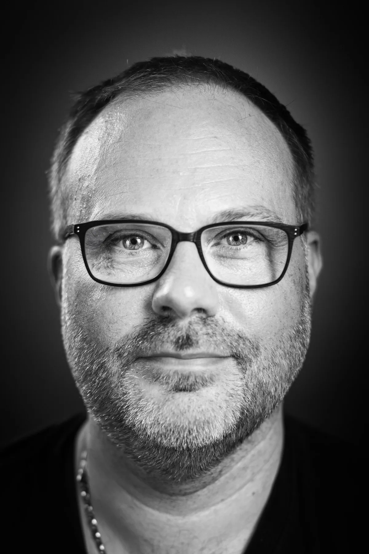 Mann mit kurzem Haar, Dreitagebart und dunkler Brille blickt ernst, dünne Kette um den Hals