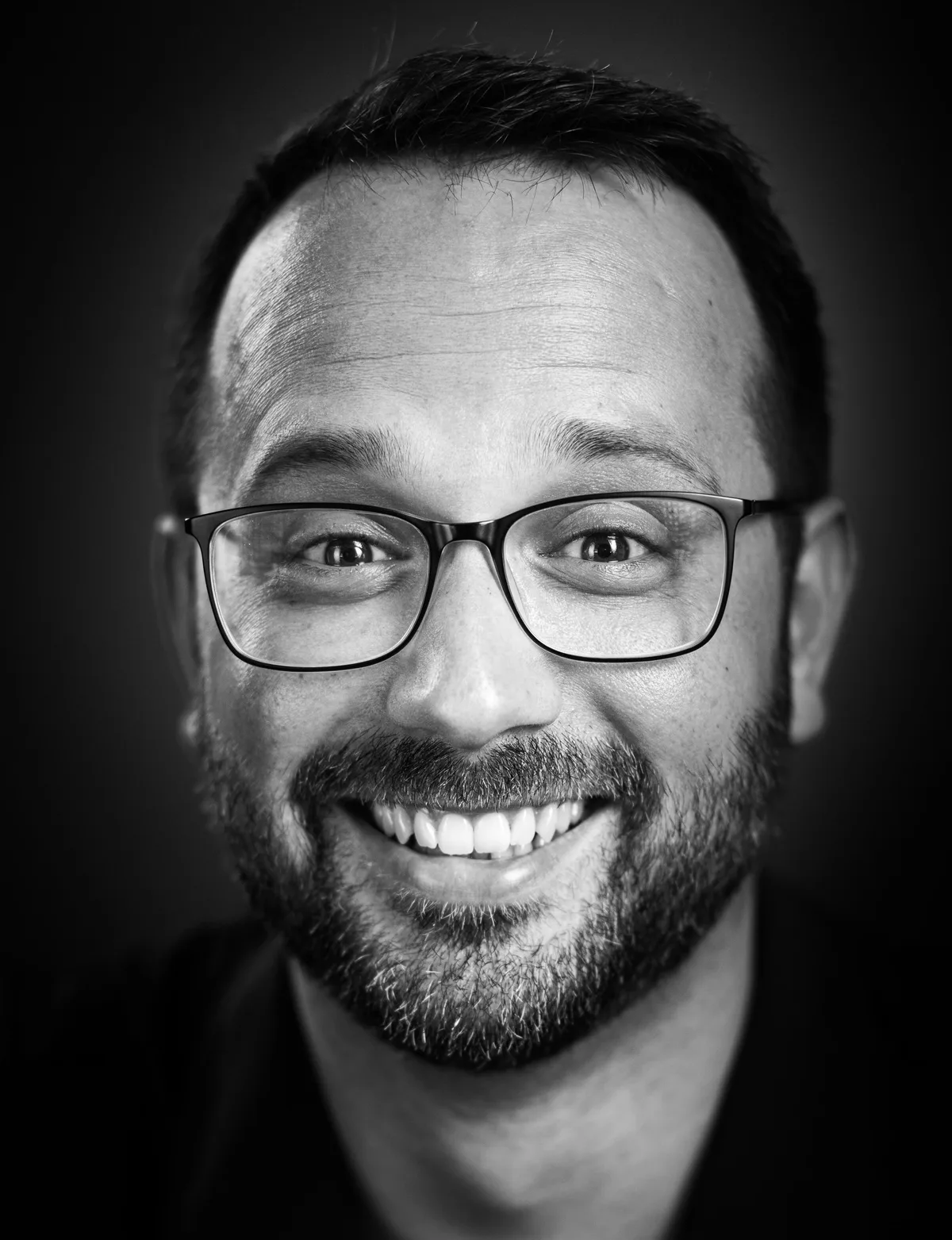 Lachender Mann mit kurzem dunklem Haar, Bart und schmaler Brille, Kopf leicht geneigt, sehr nah fotografiert