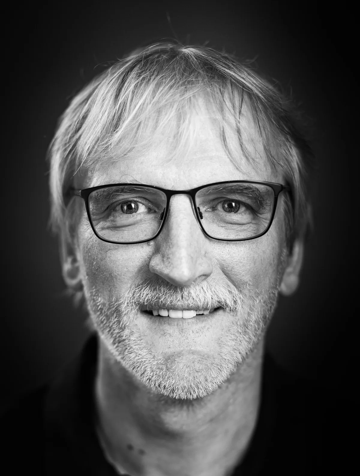 Mann mit hellem, strubbeligem Haar, grau-weißem Bart und schmaler dunkler Brille blickt freundlich geradeaus