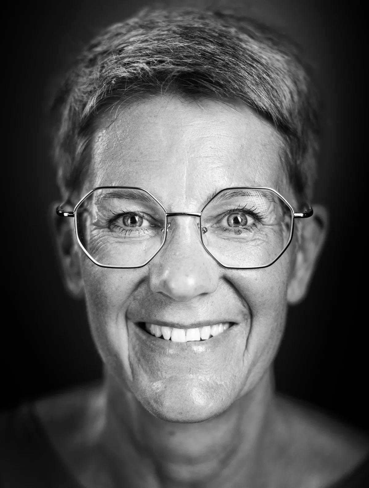 Frau mit kurzem grauem Haar und achteckiger Metallbrille lacht offen, dunkles Oberteil