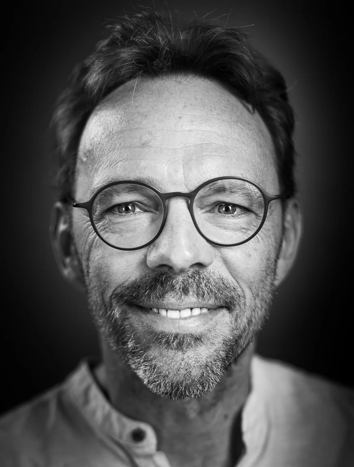Lächelnder Mann mit dunklem Haar, runder Brille und Dreitagebart, Hemd mit Stehkragen