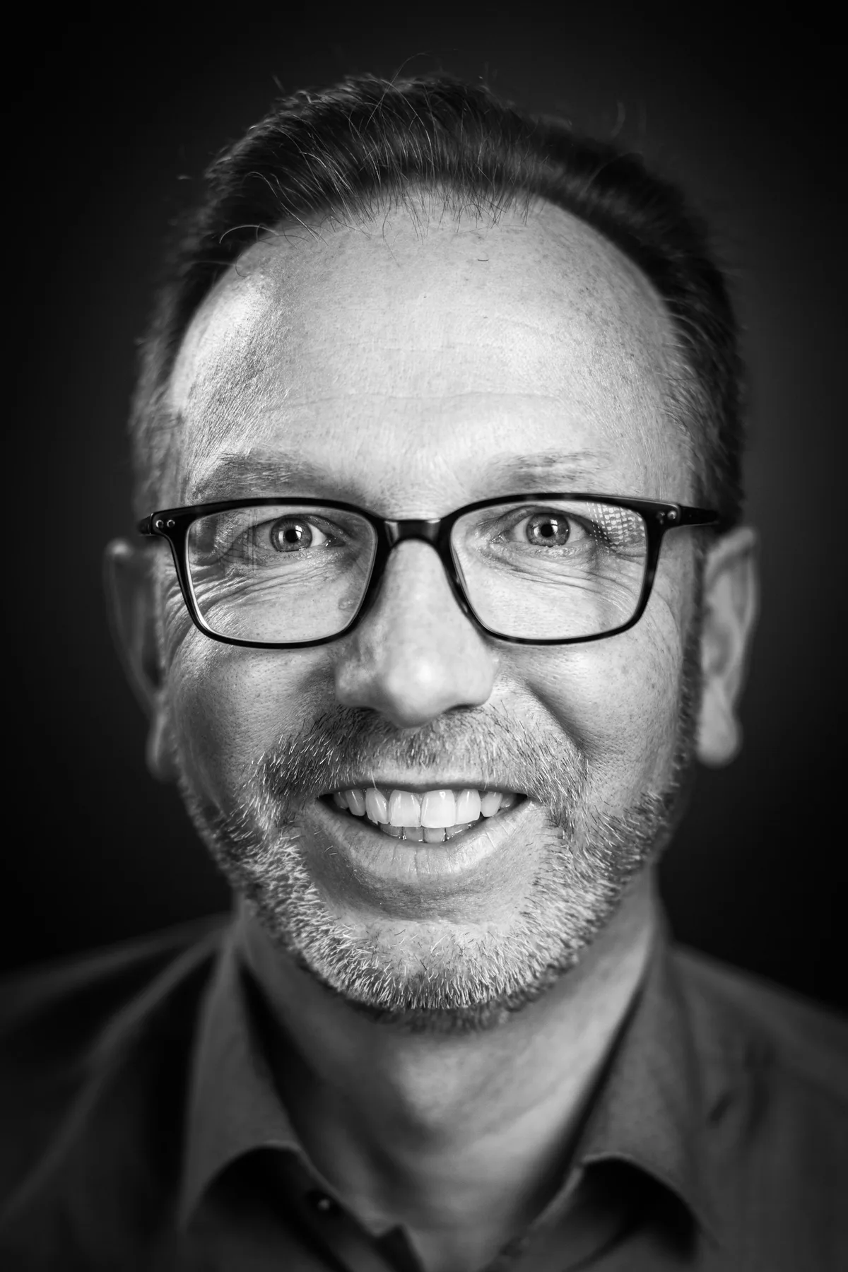 Mann mit kurzem dunklem Haar, eckiger Brille und grauem Stoppelbart lächelt, Hemd mit offenem Kragen