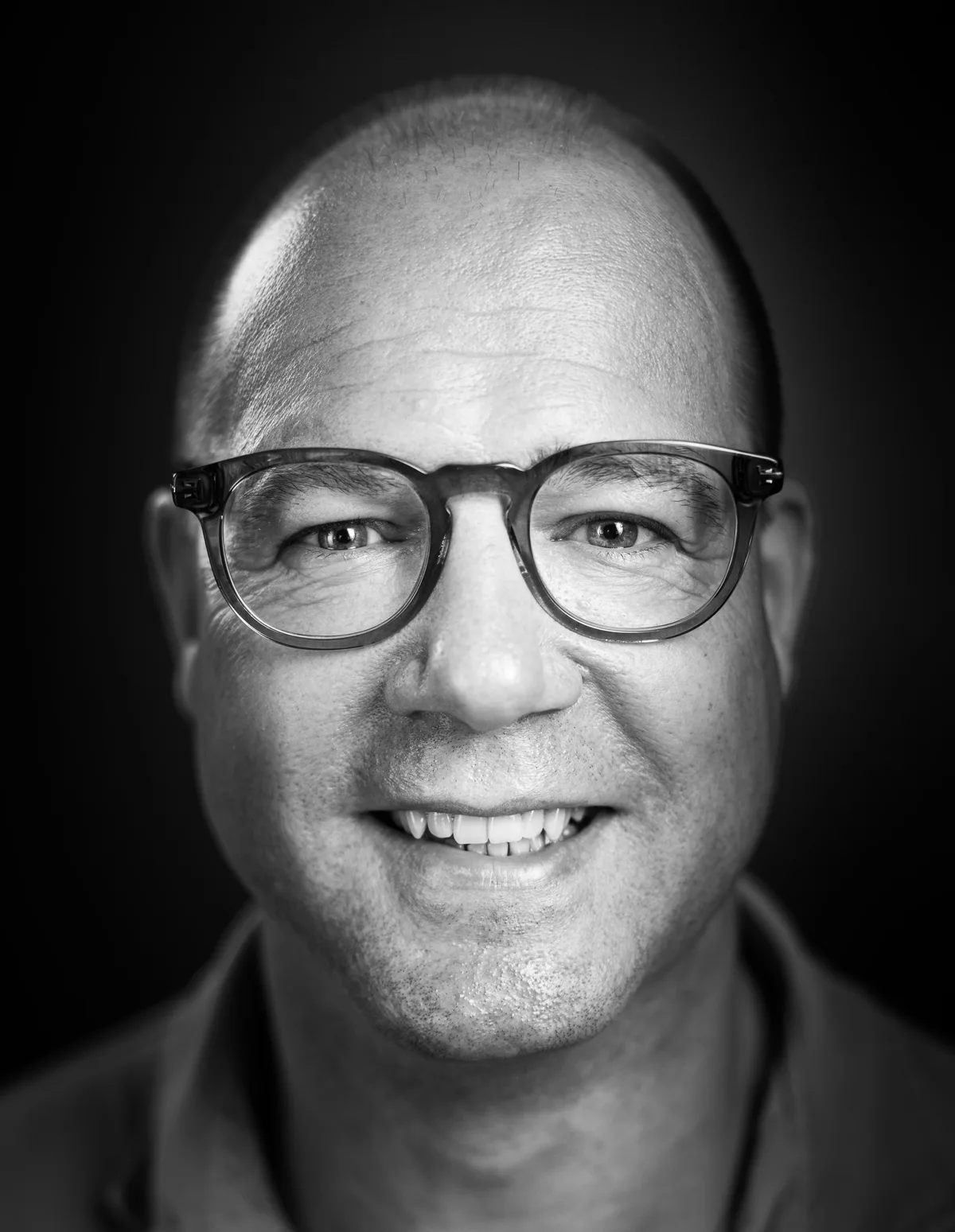 Mann mit Glatze und grau-transparenter Rundbrille lächelt schief, dunkles Polohemd
