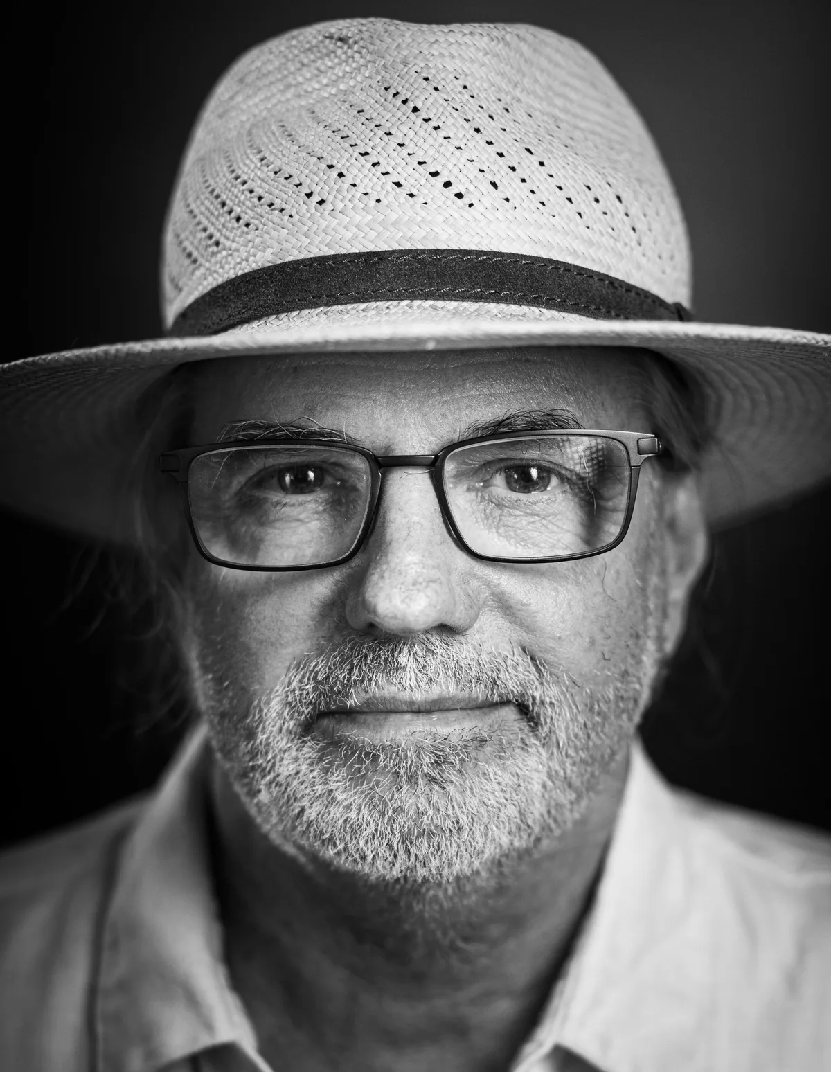 Mann mit hellem Strohhut, eckiger Brille und grau-weißem Bart blickt ernst, helles Hemd
