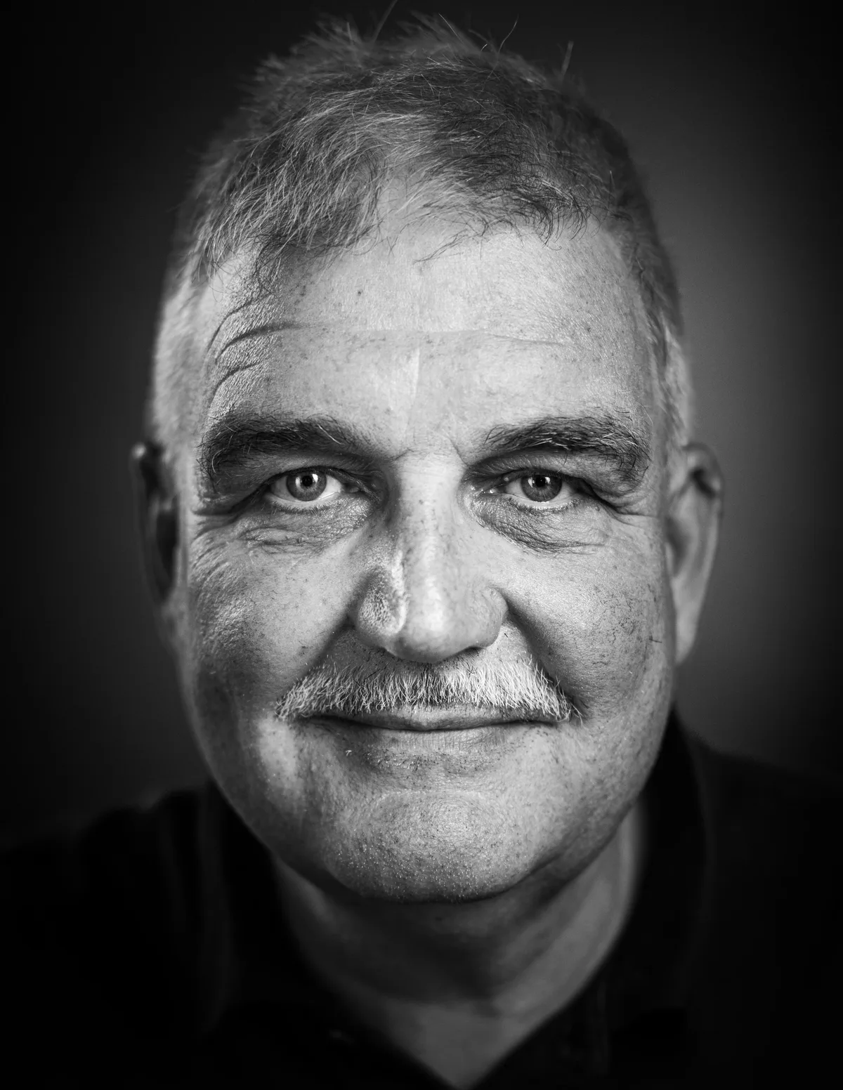 Mann mit kurzem grauem Haar und Schnauzbart blickt ruhig, die Lippen zu einem leichten Lächeln geformt