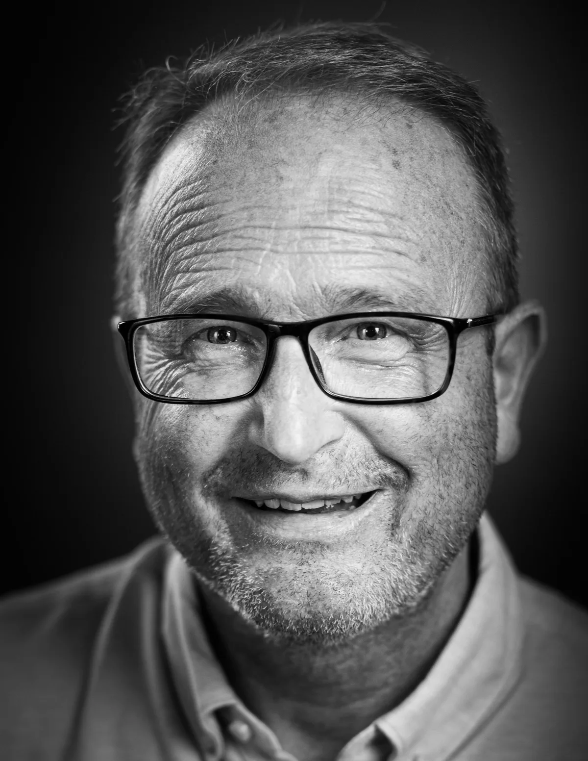 Lächelnder Mann mit schwarzer Brille, Stoppelbart, kurzem Haar und offenem Kragen, tiefe Lachfalten