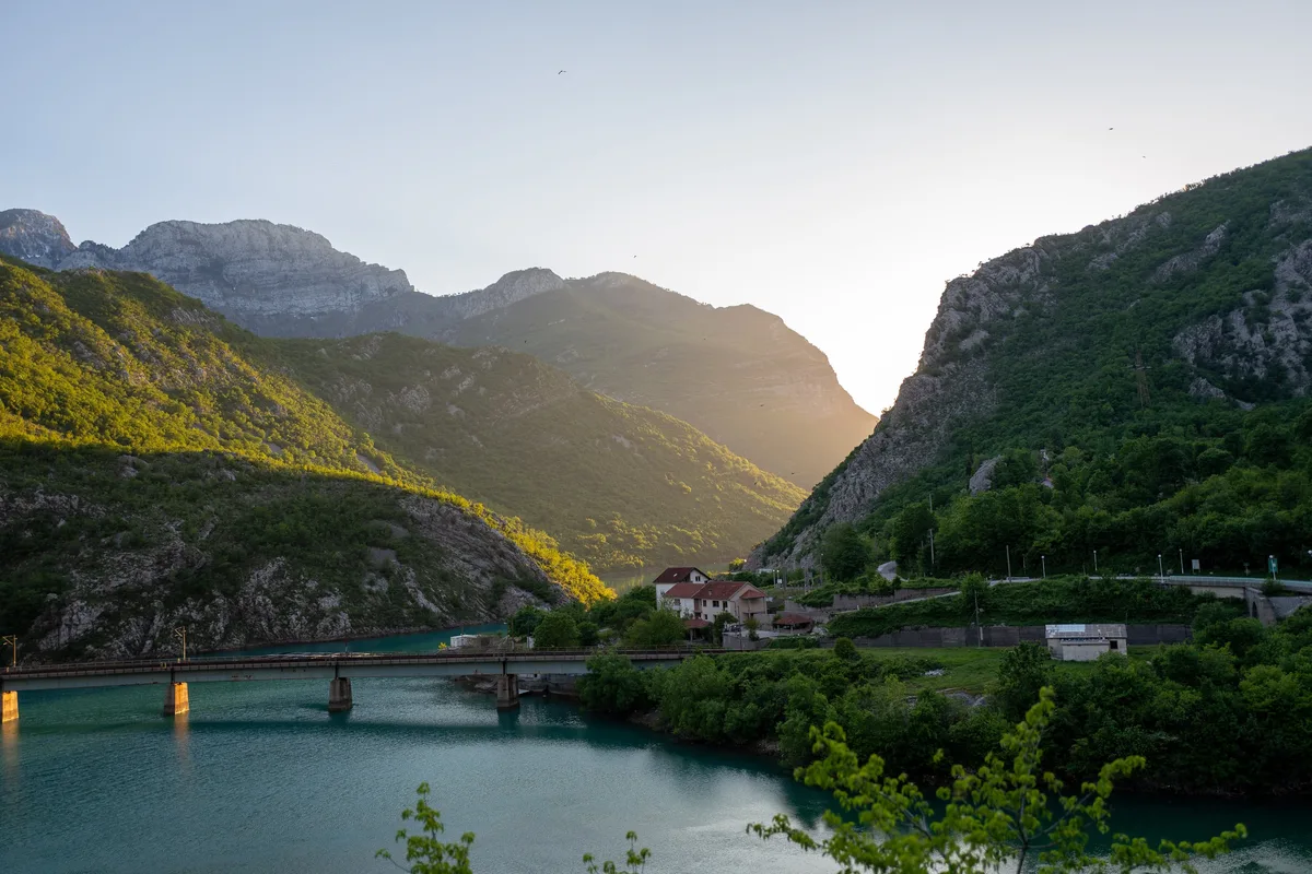 Türkisfarbener Fluss vor Bergkette bei tiefstehender Sonne, links eine Brücke, rechts ein Haus mit rotem Dach am Hang