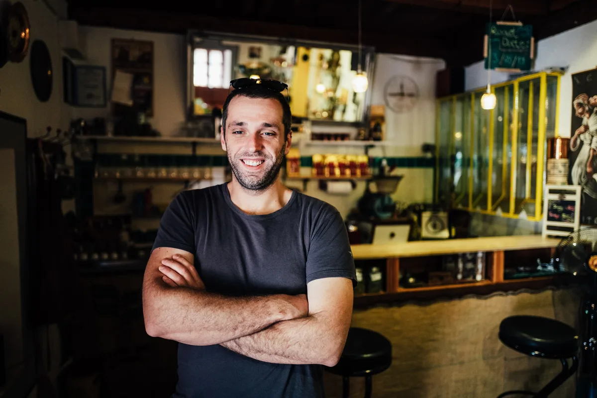 Lächelnder Mann mit verschränkten Armen und Sonnenbrille im Haar in einem kleinen Café mit Holztresen und gelber Vitrine