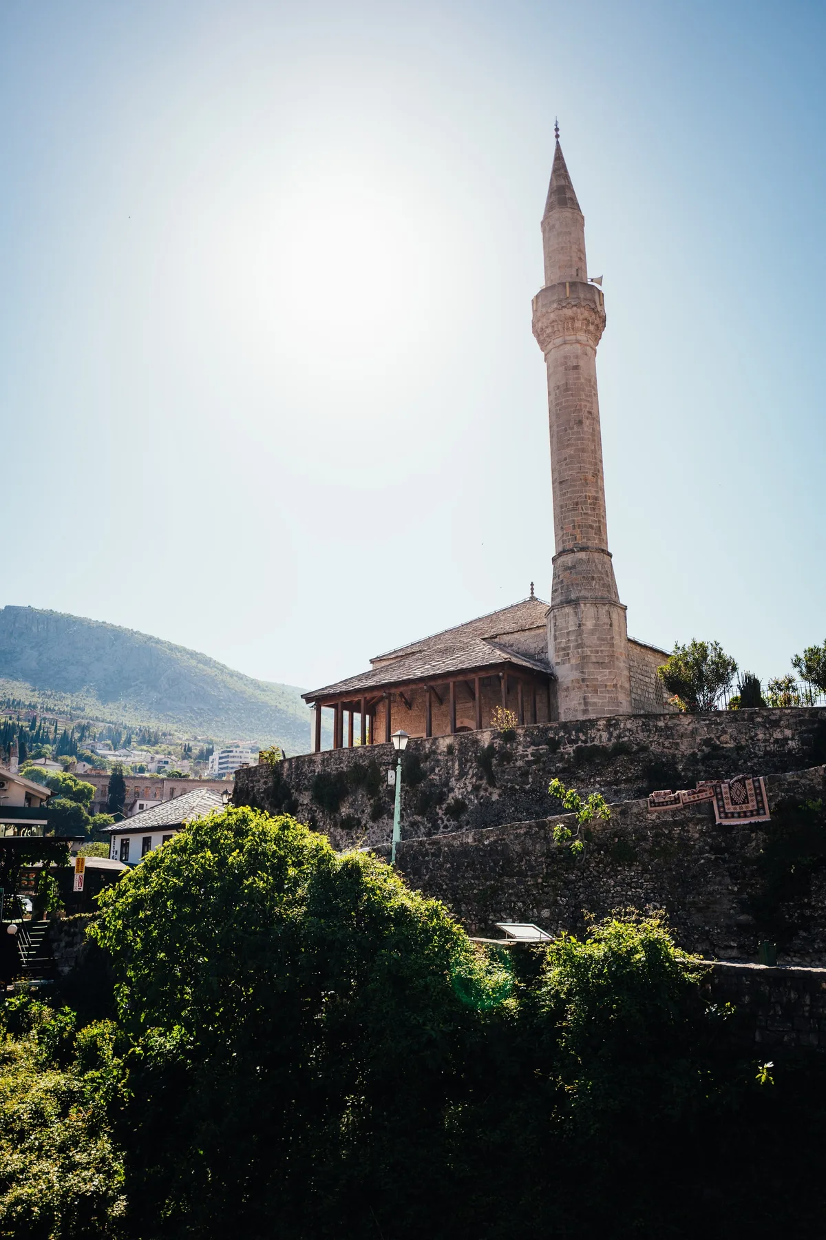 Schlankes Minarett einer Moschee mit Holzvorbau über einer Steinmauer, im Gegenlicht, Büsche im Vordergrund, Hochformat