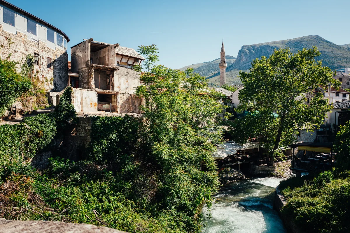 Halb verfallenes Haus am Flussufer zwischen Bäumen, in der Ferne ein Minarett vor einem kahlen Berg