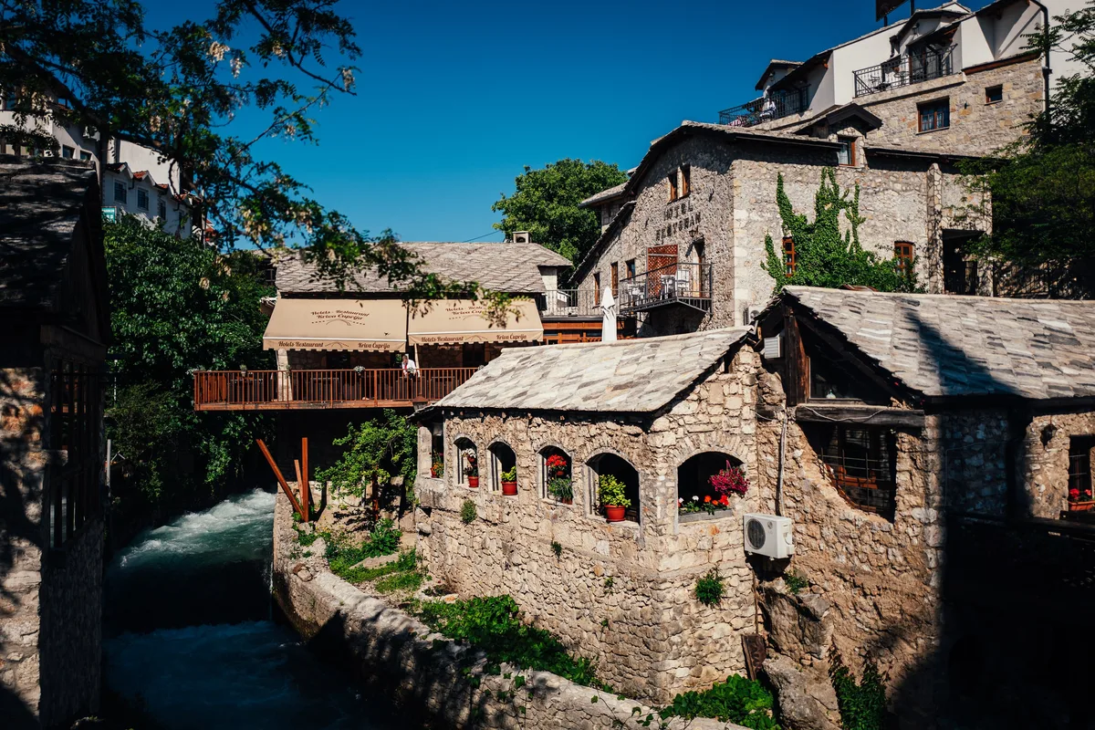 Steinhaus mit Blumenkästen in Bogenfenstern über dem Fluss, links eine Restaurantterrasse aus Holz mit beigen Markisen