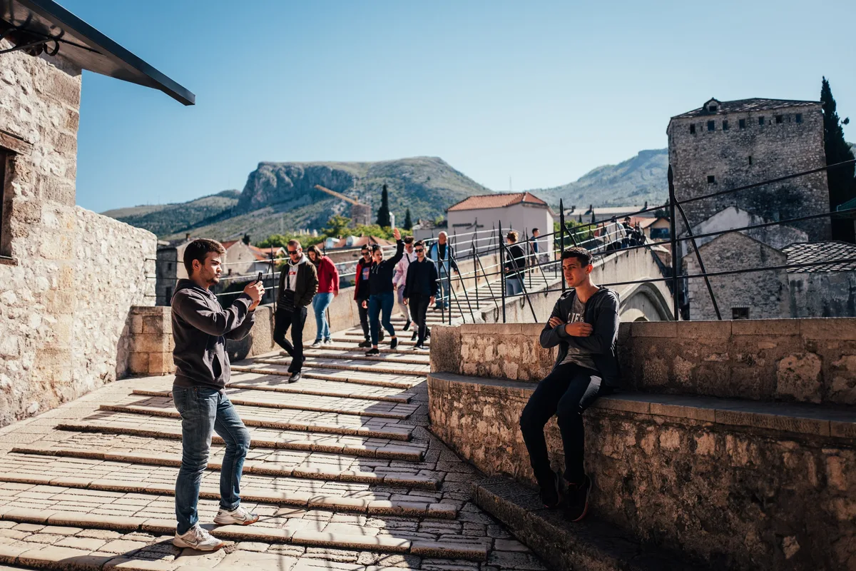 Junger Mann fotografiert mit dem Handy auf einer Steintreppe, ein zweiter sitzt auf der Mauer, dahinter Passanten