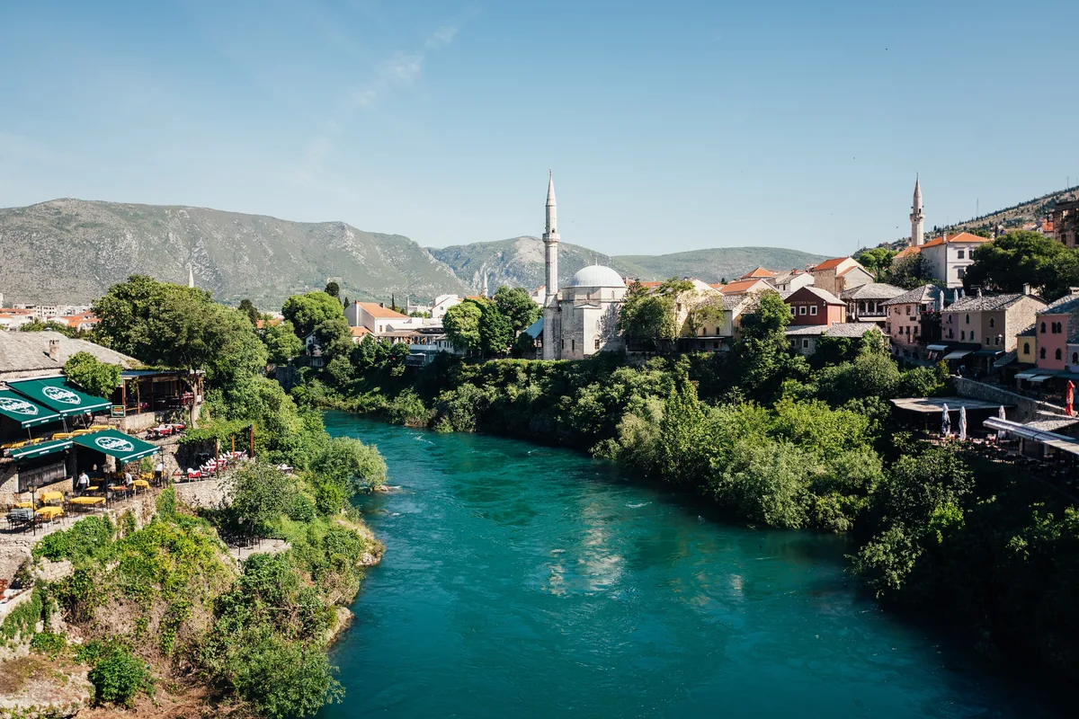 Türkisgrüner Fluss zwischen Grünstreifen, am Ufer eine Moschee mit Kuppel und Minarett sowie Häuser mit roten Dächern