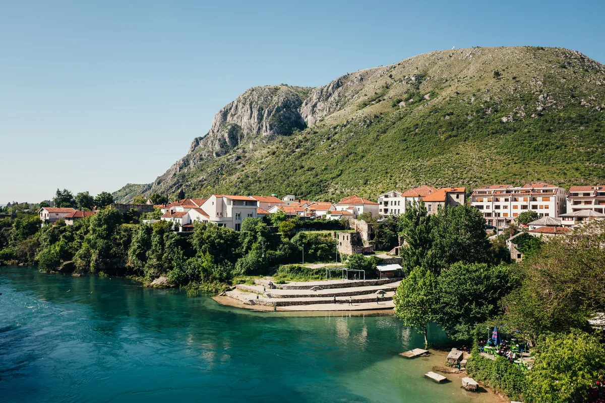 Türkisfarbener Fluss mit Stufen am Ufer, dahinter Häuser mit roten Dächern vor einem steilen, felsigen Berghang