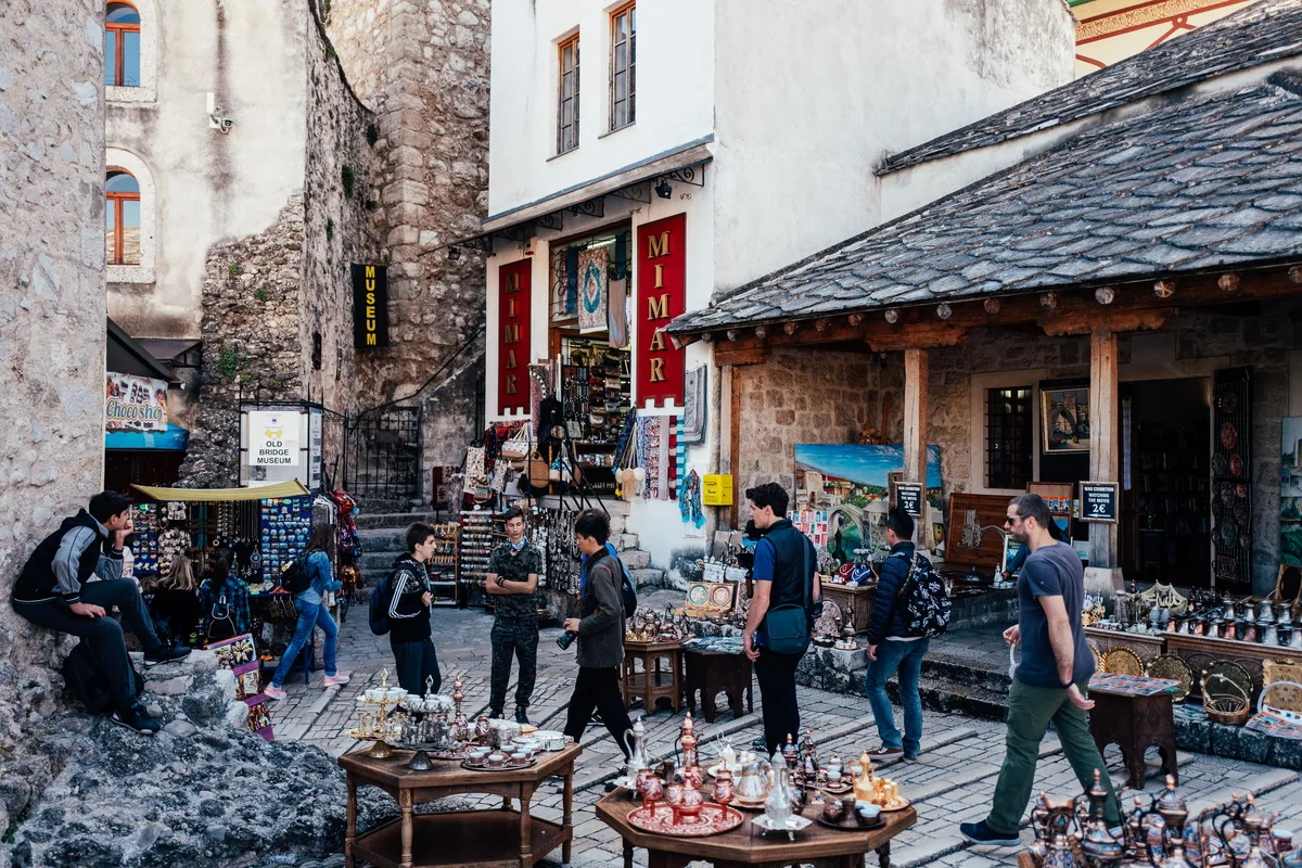 Souvenirstände mit Kupfergeschirr in einer Altstadtgasse, davor Touristen mit Rucksäcken, rechts ein Mann in Cargohose