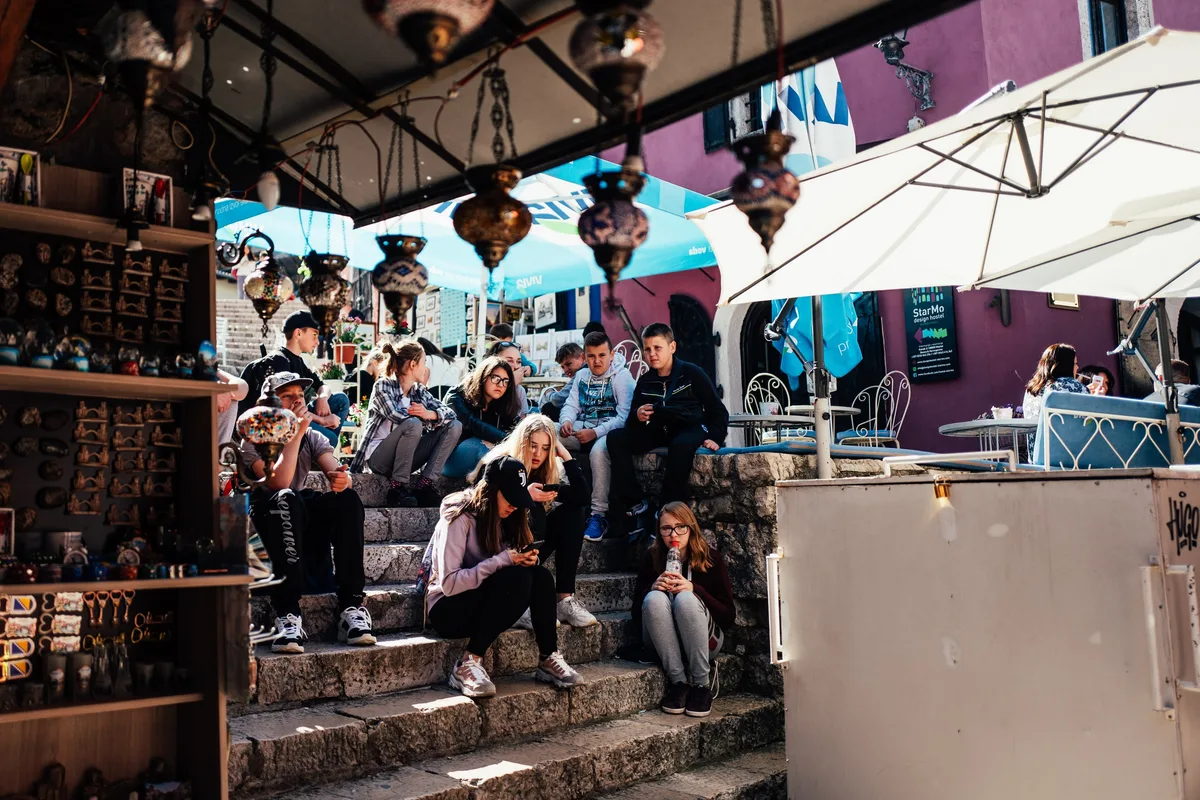 Jugendliche sitzen mit Handys auf einer Steintreppe zwischen Souvenirregal und Café, darüber hängen Laternen