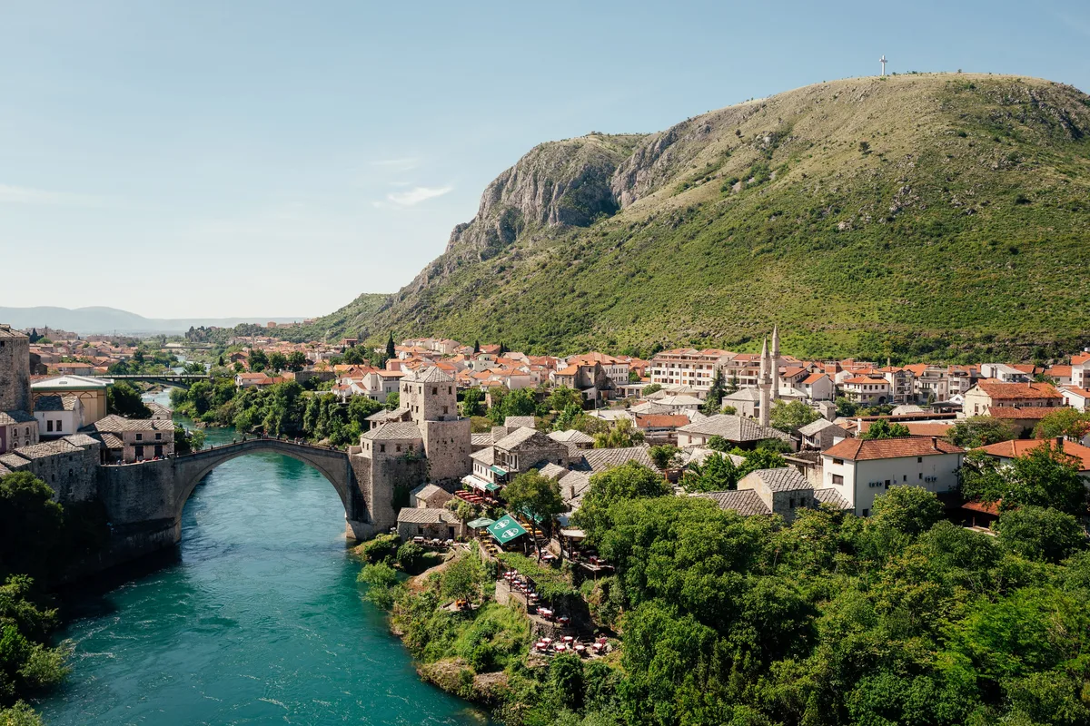 Blick von oben auf Fluss und steinerne Bogenbrücke, Altstadt mit roten Dächern und Minaretten, dahinter ein Berg mit Kreuz