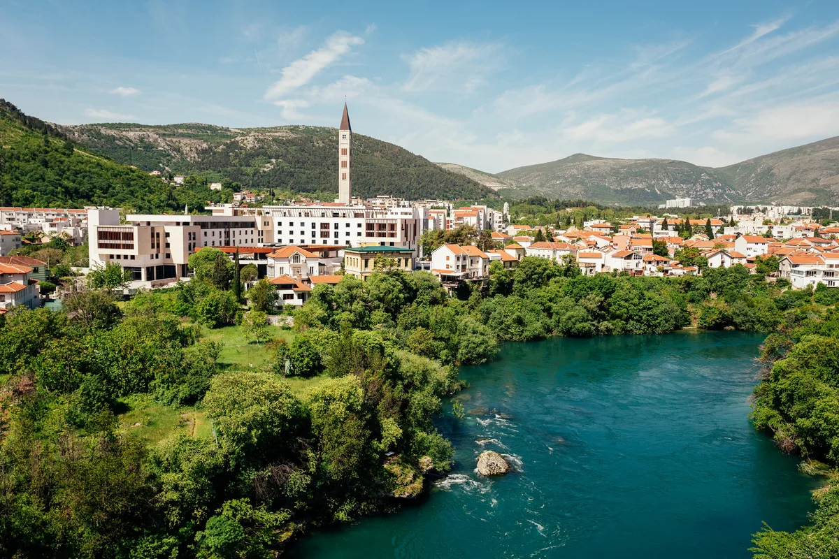 Stadt am Fluss mit schlankem Kirchturm und roten Dächern, im Vordergrund türkisfarbenes Wasser mit Stromschnellen