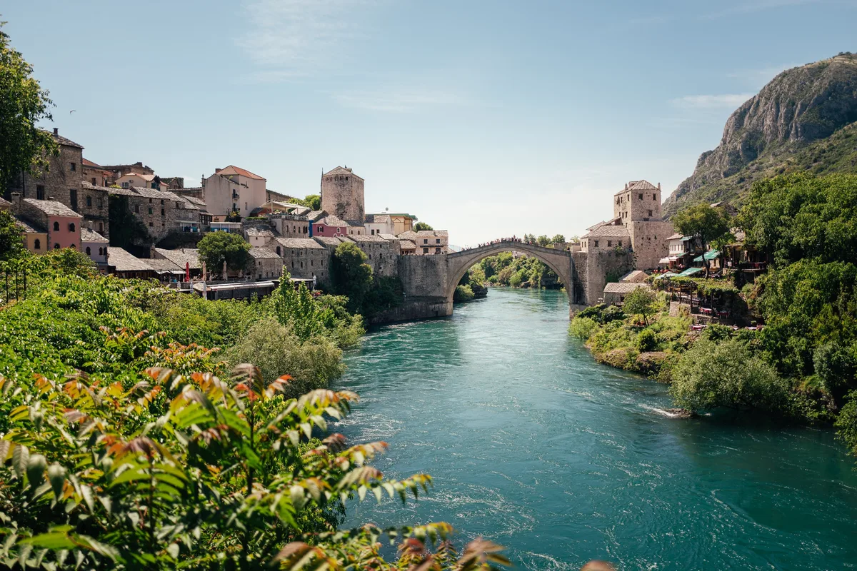 Türkisfarbener Fluss mit steinerner Bogenbrücke, links Häuser am Hang, rechts Felsen, vorn Laub mit rötlichen Blättern