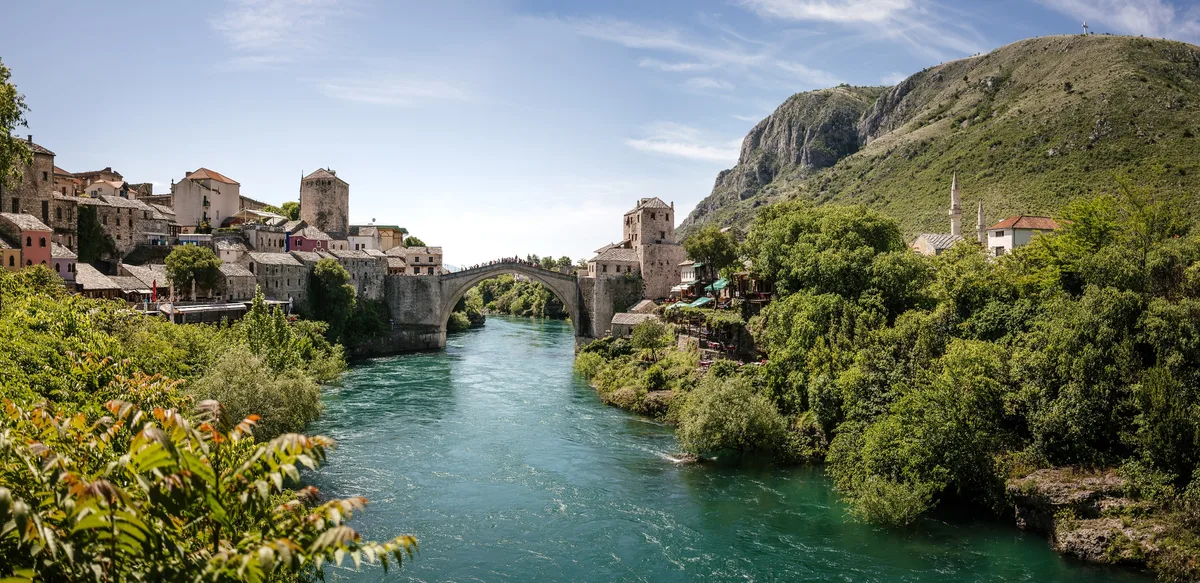 Panorama mit Fluss, Bogenbrücke und Altstadthäusern, rechts bewachsene Uferböschung und ein steil aufragender Berg