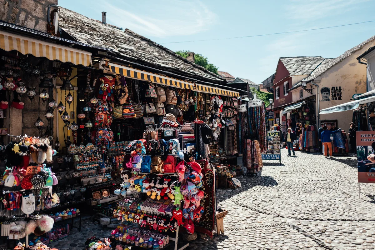 Gasse mit Souvenirständen, Stofftieren und Laternen unter gestreiften Markisen, rechts Kopfsteinpflaster und Passanten