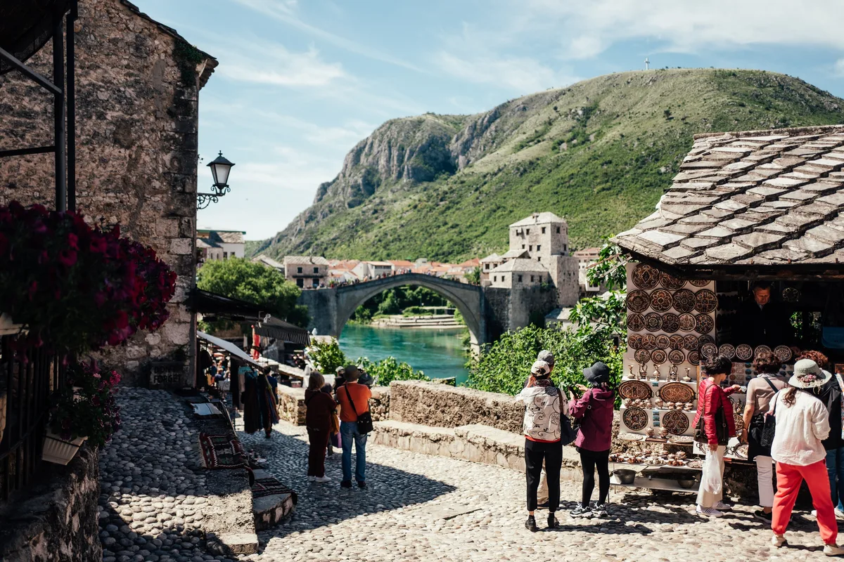 Touristen stehen auf einer Pflastergasse mit Blick auf eine ferne Bogenbrücke, rechts ein Stand mit Kupferplatten