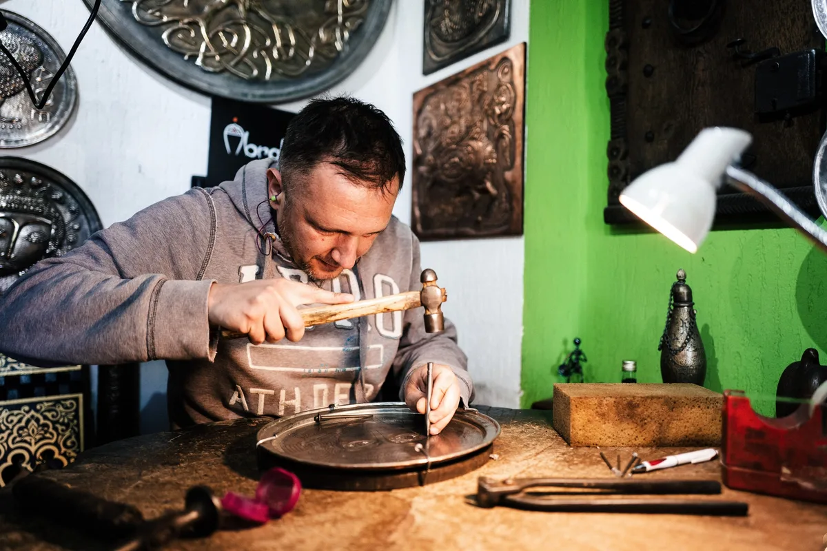 Mann in grauem Kapuzenpulli treibt mit einem Hammer ein Muster in eine Kupferplatte, hinter ihm Metallteller an der Wand