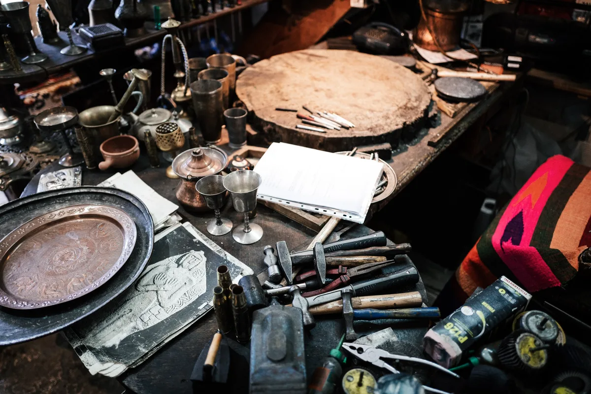Werkbank voller Hämmer, Zangen, Kupferkannen und Metallbecher, links ein Tablett mit Ornamenten, hinten ein runder Holzblock