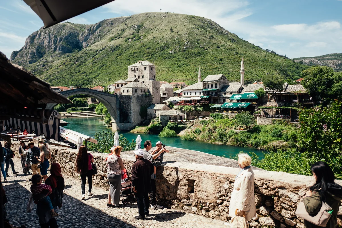 Touristen stehen und fotografieren auf einer Steinmauer an der Gasse, dahinter Fluss, Bogenbrücke und Häuser vor einem Berg