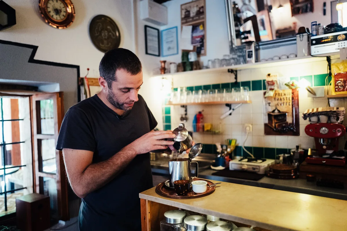 Mann in schwarzem T-Shirt gießt aus einer kleinen Kanne Kaffee in ein Gefäß, rechts eine rote Espressomaschine