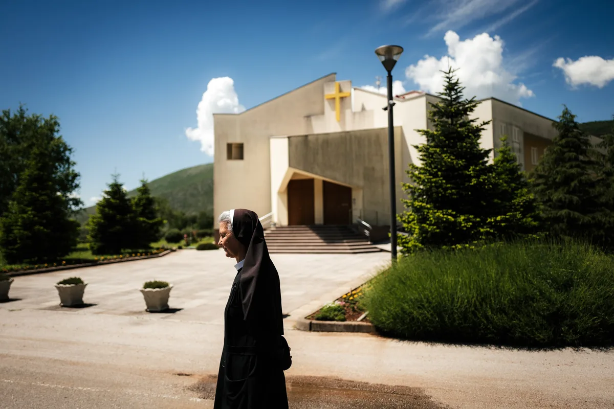 Ordensfrau in schwarzem Habit geht im Profil an einem Platz vorbei, dahinter eine Kirche mit goldenem Kreuz und Tannen
