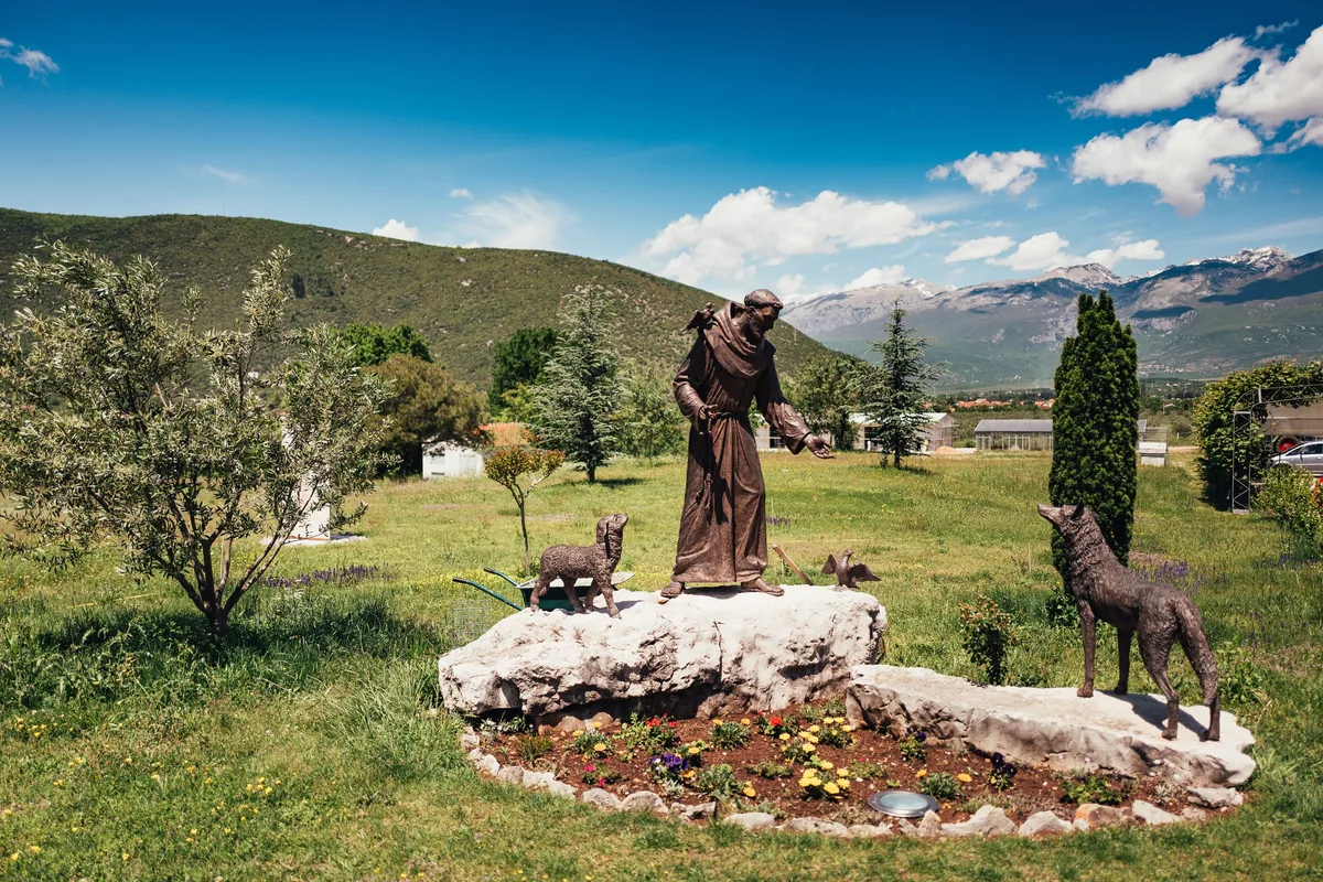 Bronzefigur eines Mönchs mit Vogel auf der Schulter auf einem Felsblock, davor zwei Tierfiguren, dahinter Wiese und Berge
