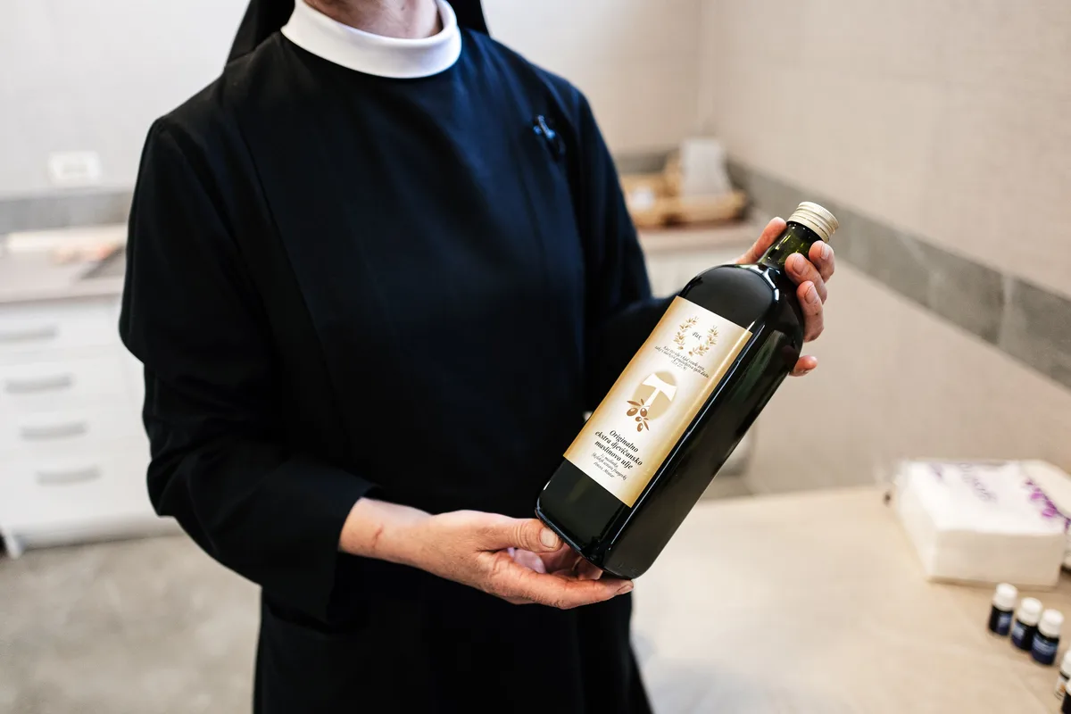 Ordensfrau in schwarzem Habit hält eine Flasche Olivenöl mit Etikett in beiden Händen, rechts auf dem Tisch kleine Fläschchen