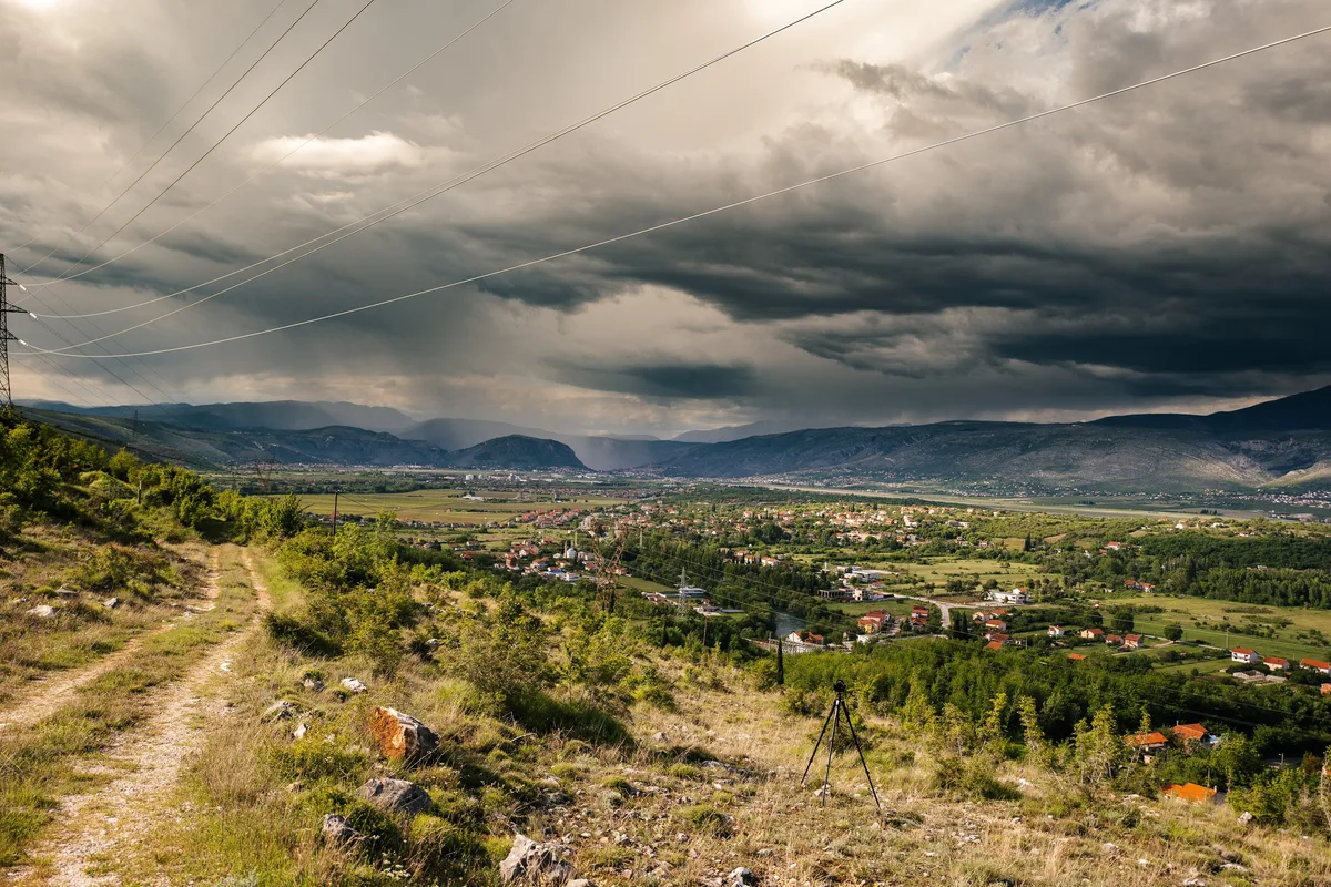 Dunkle Gewitterwolken über einem Tal mit verstreuten Häusern, im Vordergrund ein Feldweg und ein Stativ am Hang