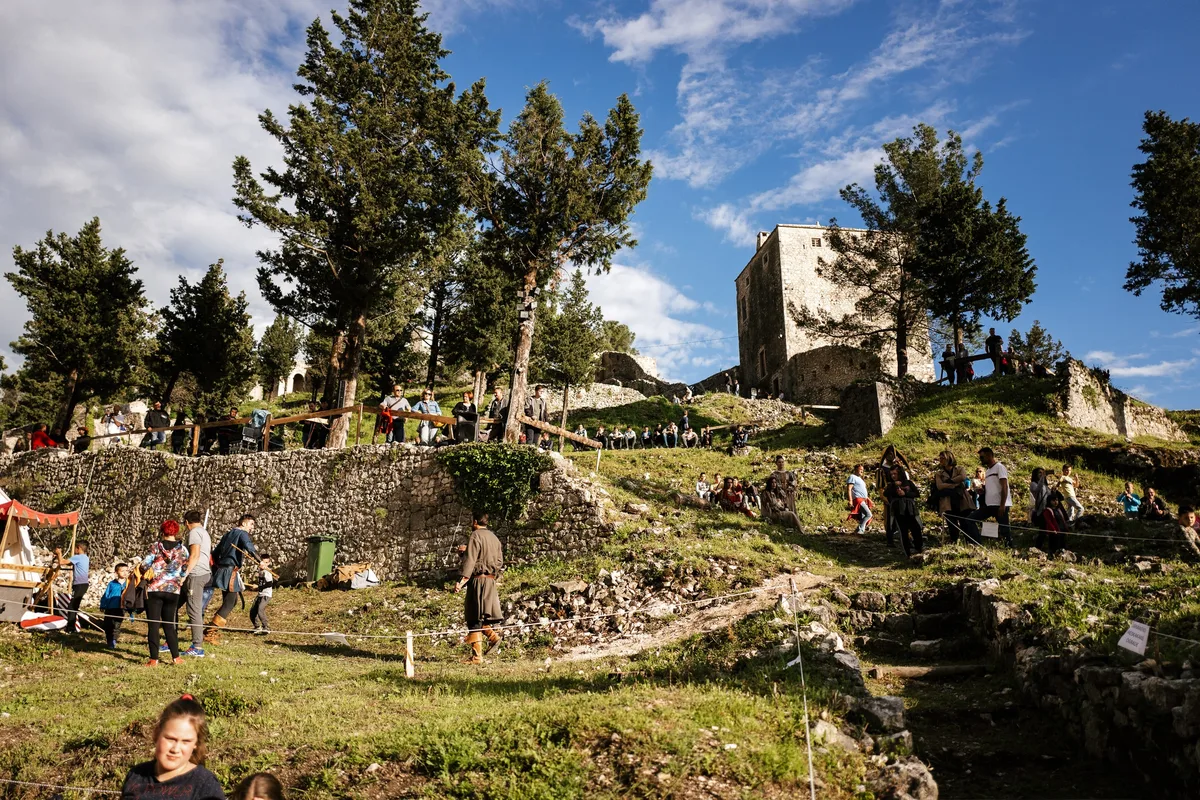 Schräger Blick auf einen Hang mit Burgturm, Mauern und Zuschauern, vorn ein Mann in brauner Tunika und spielende Kinder