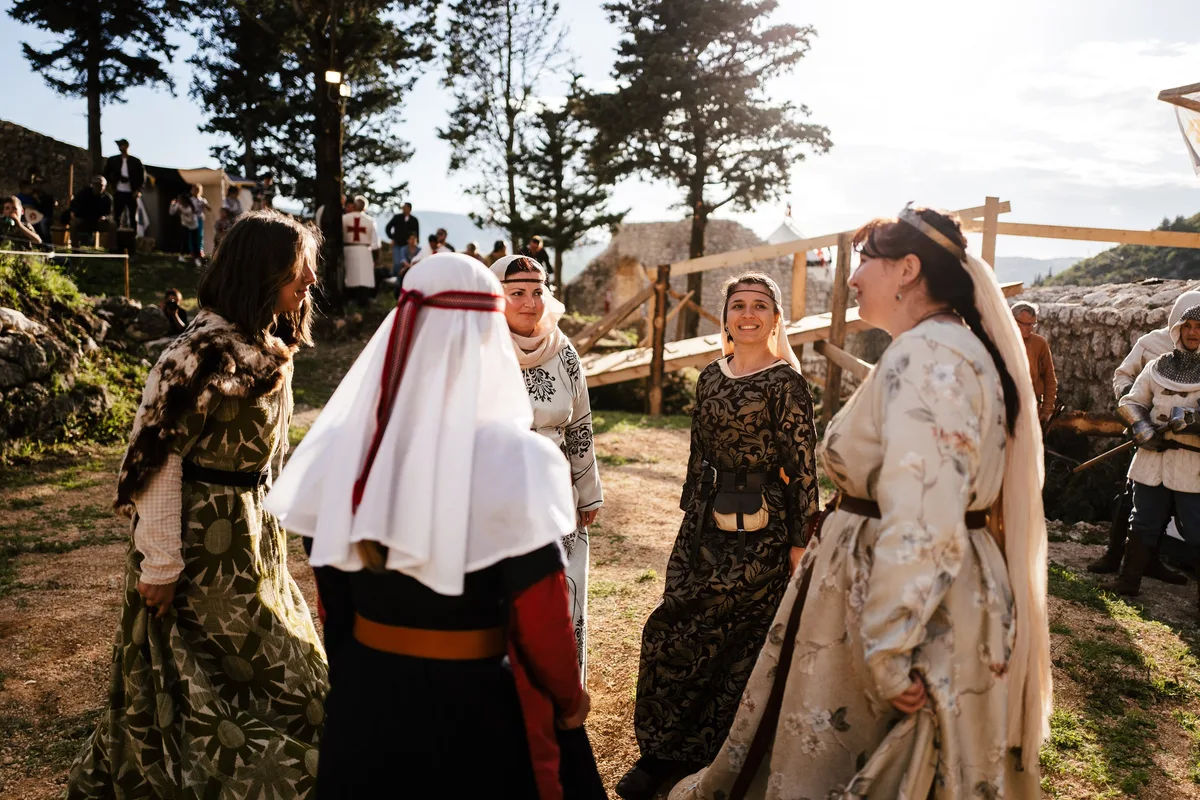 Frauen in Gewändern stehen lachend im Kreis, links Fellweste, in der Mitte eine Frau mit Stirnband, rechts beiges Kleid
