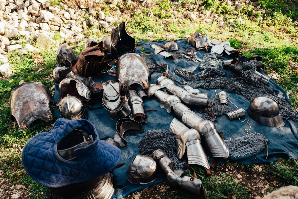 Metallene Rüstungsteile, Kettenhemd und Helme liegen auf einer blauen Decke im Gras, links eine wattierte Haube