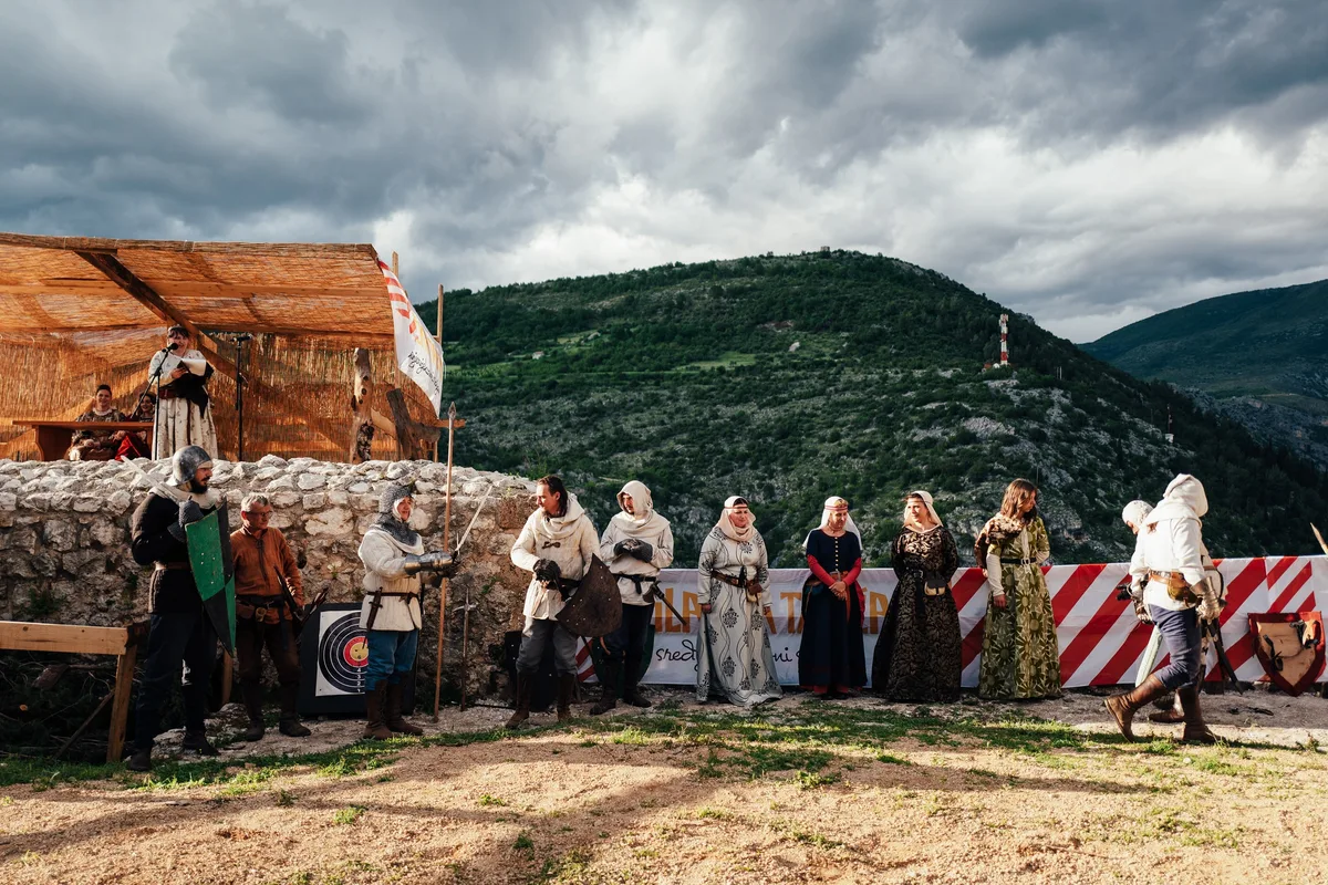 Frauen in Gewändern und Männer in Rüstung stehen in einer Reihe vor Strohdachtribüne und Absperrband unter dunklen Wolken