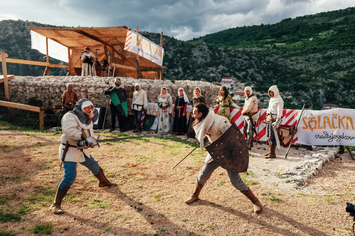 Zwei Schwertkämpfer mit Holzschild und Kettenhaube umkreisen sich auf Sandboden, hinter ihnen Zuschauer und ein Festbanner