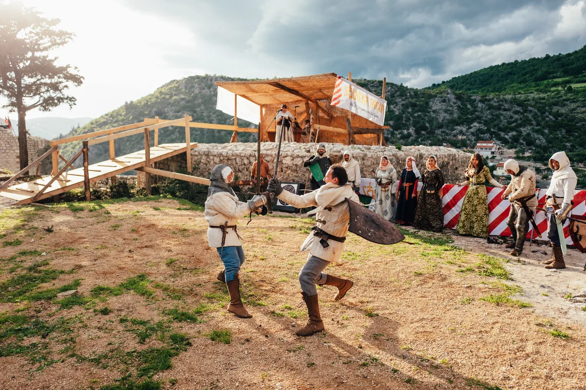 Zwei Kämpfer mit Schwertern im Gegenlicht auf Sandboden, links ein Holzsteg, rechts Zuschauerinnen vor dem Absperrband