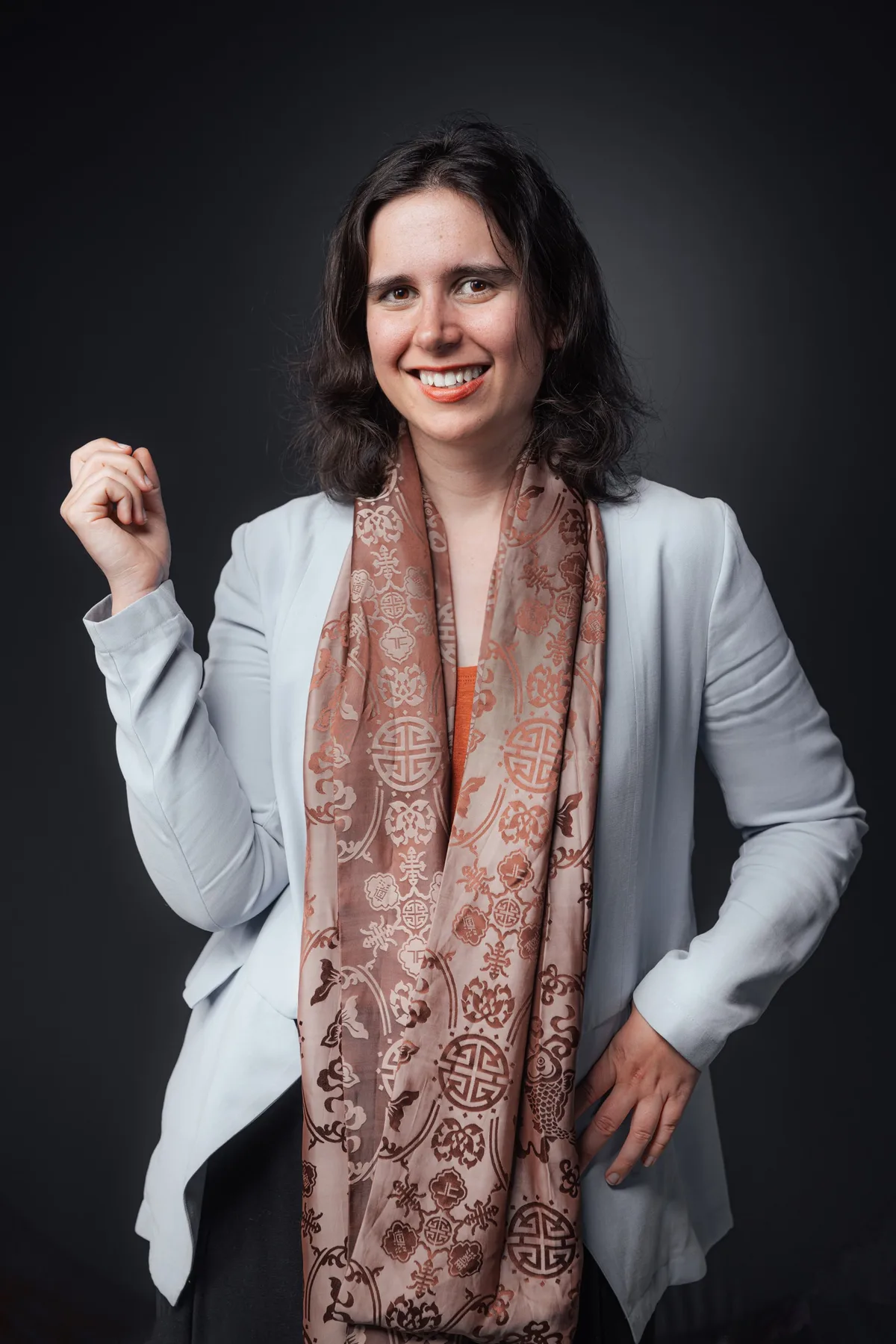 Lachende junge Frau in hellem Blazer mit orangem Seidenschal mit Ornamenten, eine Faust erhoben, die andere Hand in der Hüfte