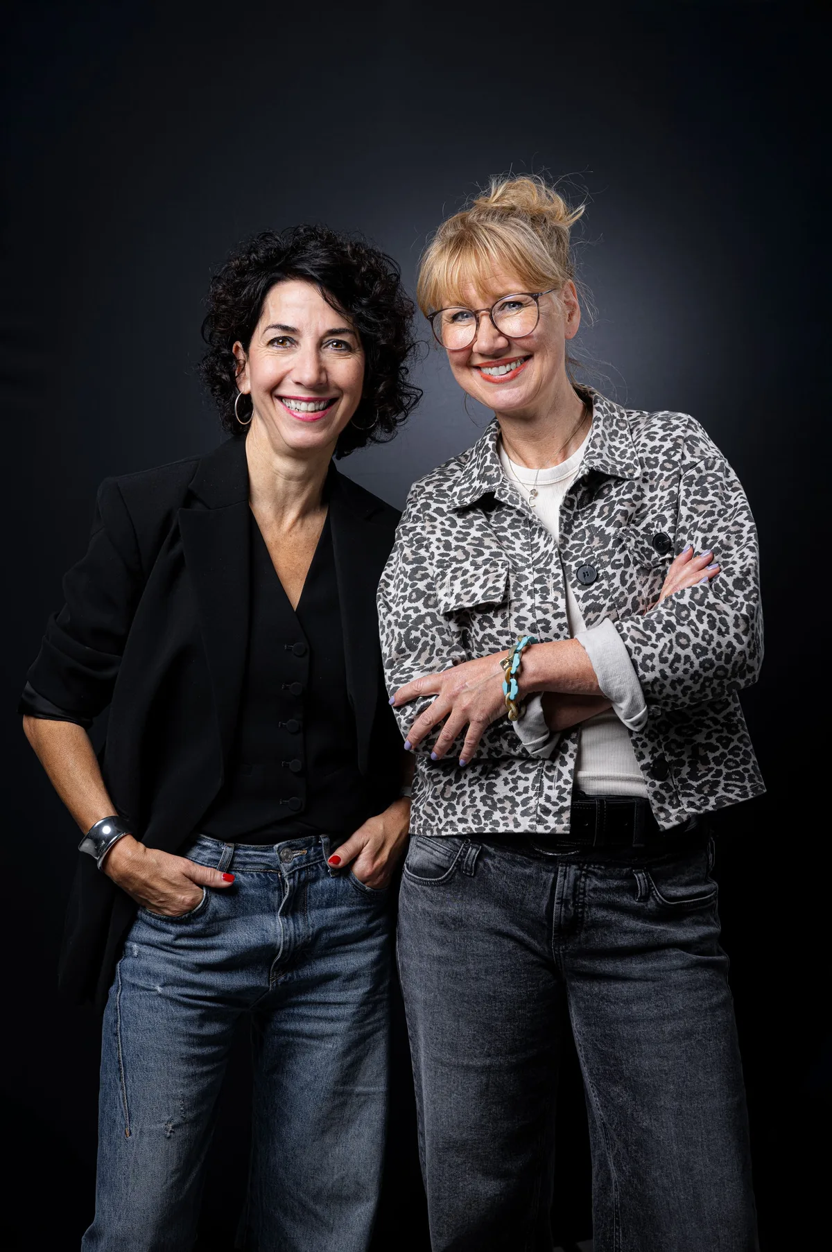 Zwei lachende Frauen nebeneinander, links mit schwarzen Locken in Schwarz und Jeans, rechts mit Brille im Leopardenjackett