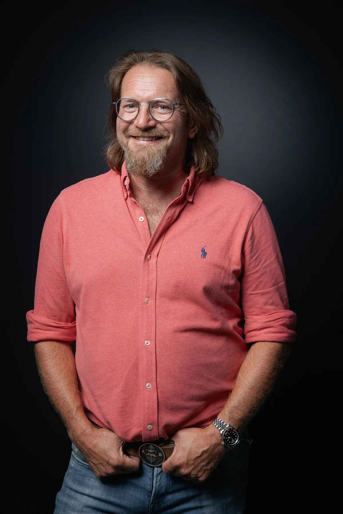 Lächelnder Mann mit langen Haaren, Brille und Bart in lachsfarbenem Polohemd, Daumen in den Hosentaschen eingehakt