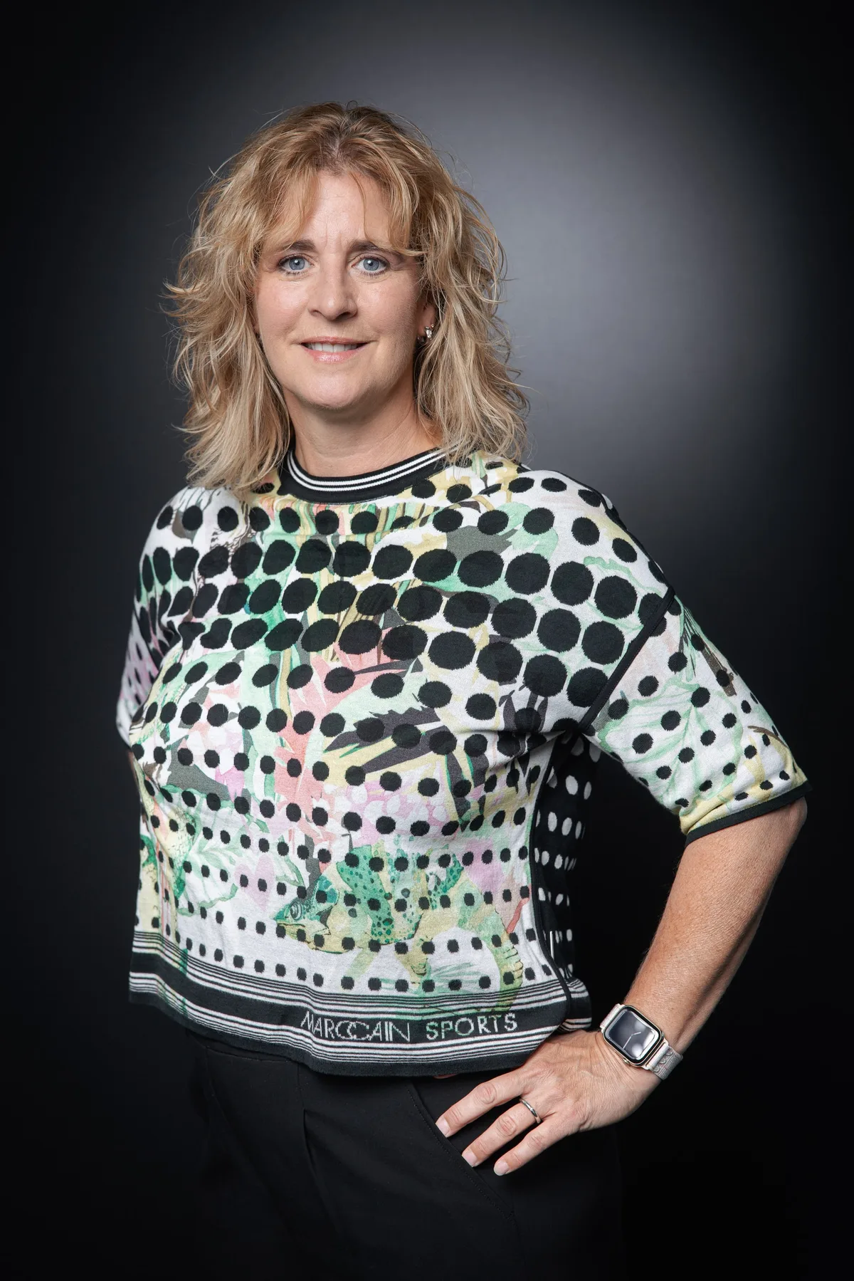 Frau mit blonden Locken in gepunktetem Shirt mit Dschungelmotiv, eine Hand in der Hüfte, ruhiger Blick in die Kamera