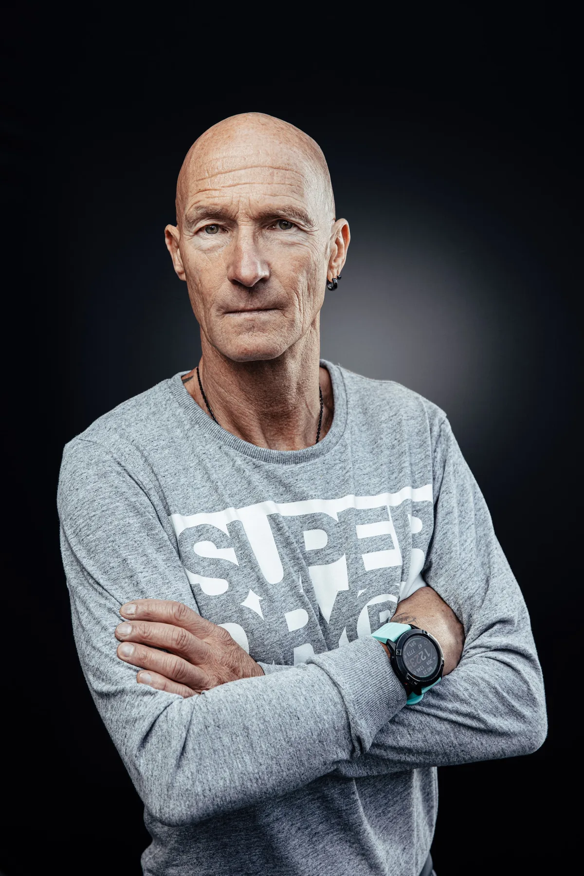 Glatzköpfiger Mann im grauen Sweatshirt mit Schriftzug, Sportuhr am Handgelenk, Arme verschränkt, konzentrierter Blick