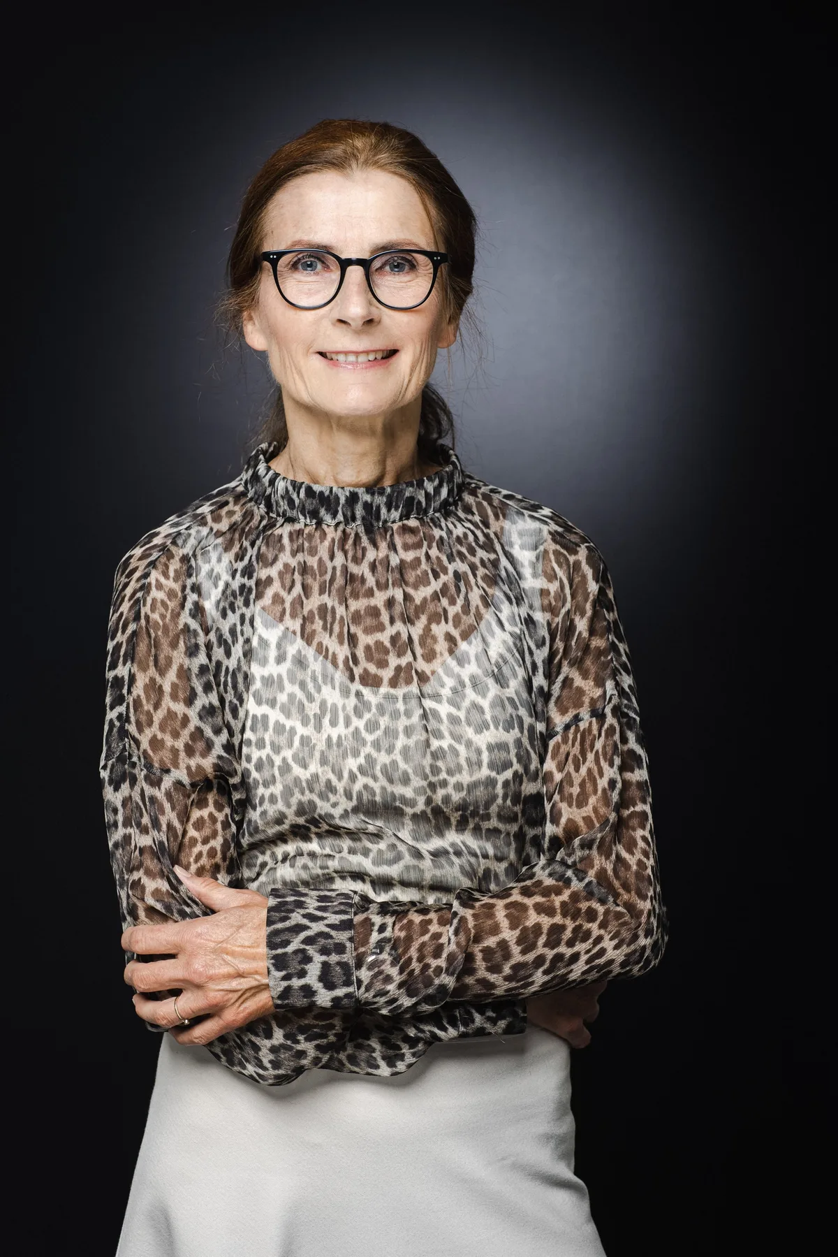 Frau mit Brille und zurückgebundenem Haar in transparenter Bluse mit Leopardenmuster, Hände vor dem Körper, leichtes Lächeln