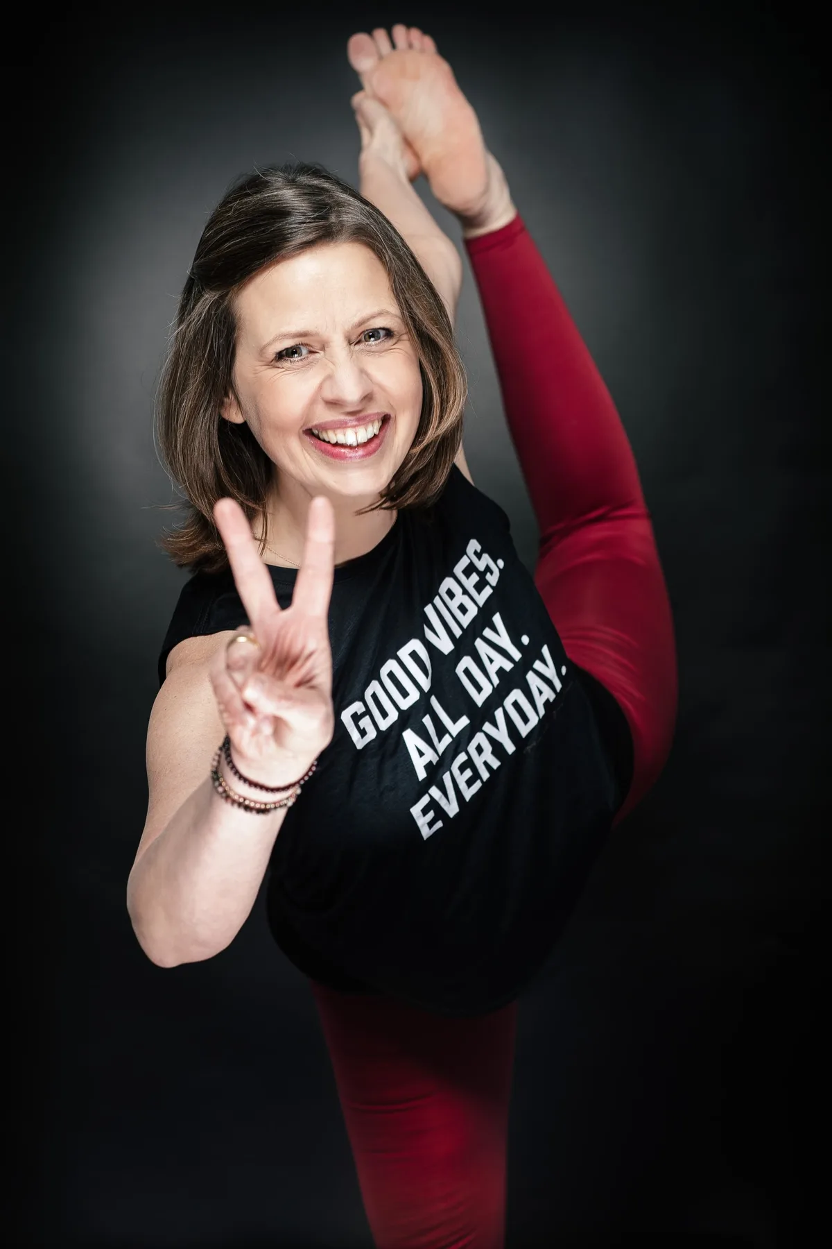 Lachende Frau im schwarzen Shirt mit Schriftzug hebt ein Bein senkrecht nach oben und zeigt mit einer Hand das Peace-Zeichen
