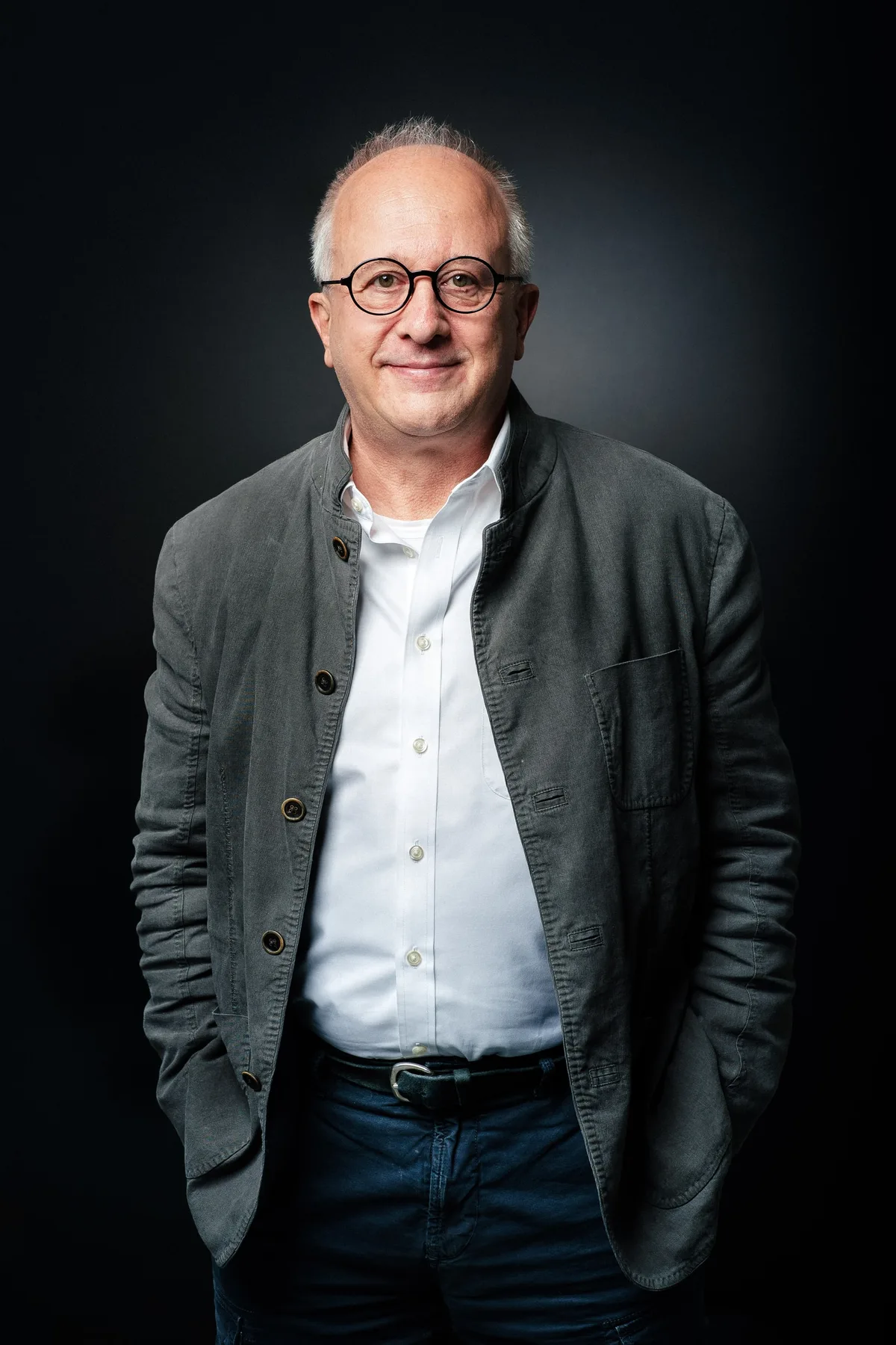 Mann mit runder Brille in grauer Leinenjacke und weißem Hemd, die Hände in den Hosentaschen, ruhiges Lächeln, Hochformat